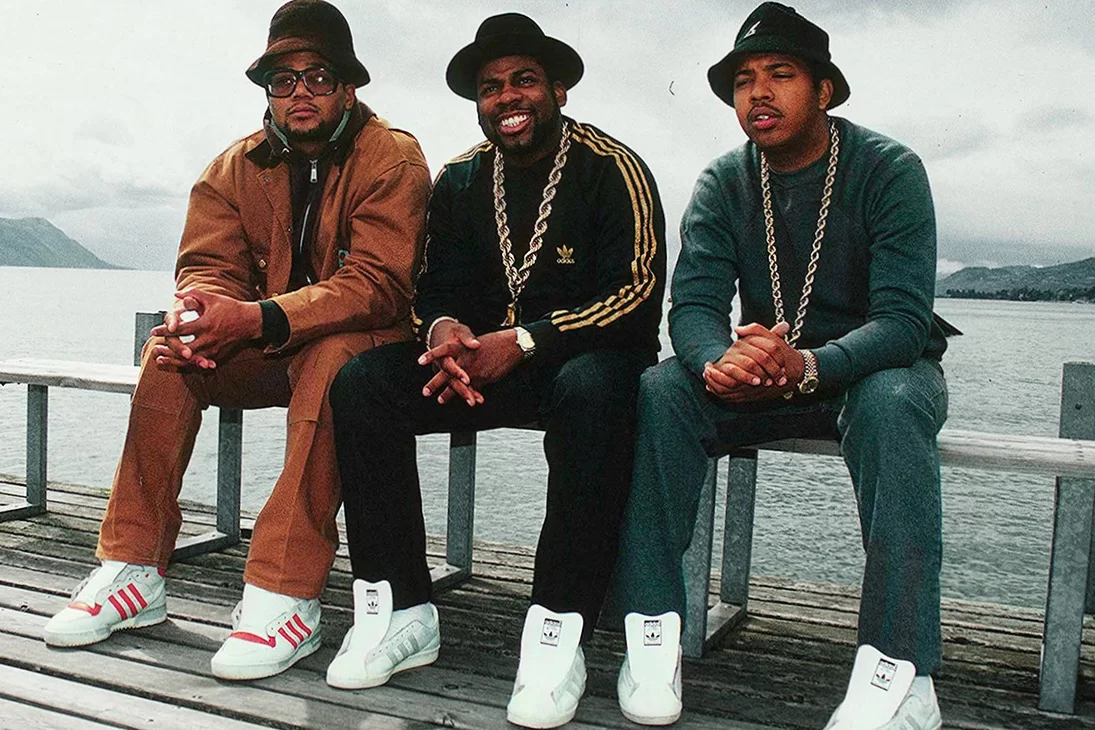 Three men dressed in iconic hip-hop fashion with tracksuits and hats sit by the waterside, embodying music's influence on fashion.